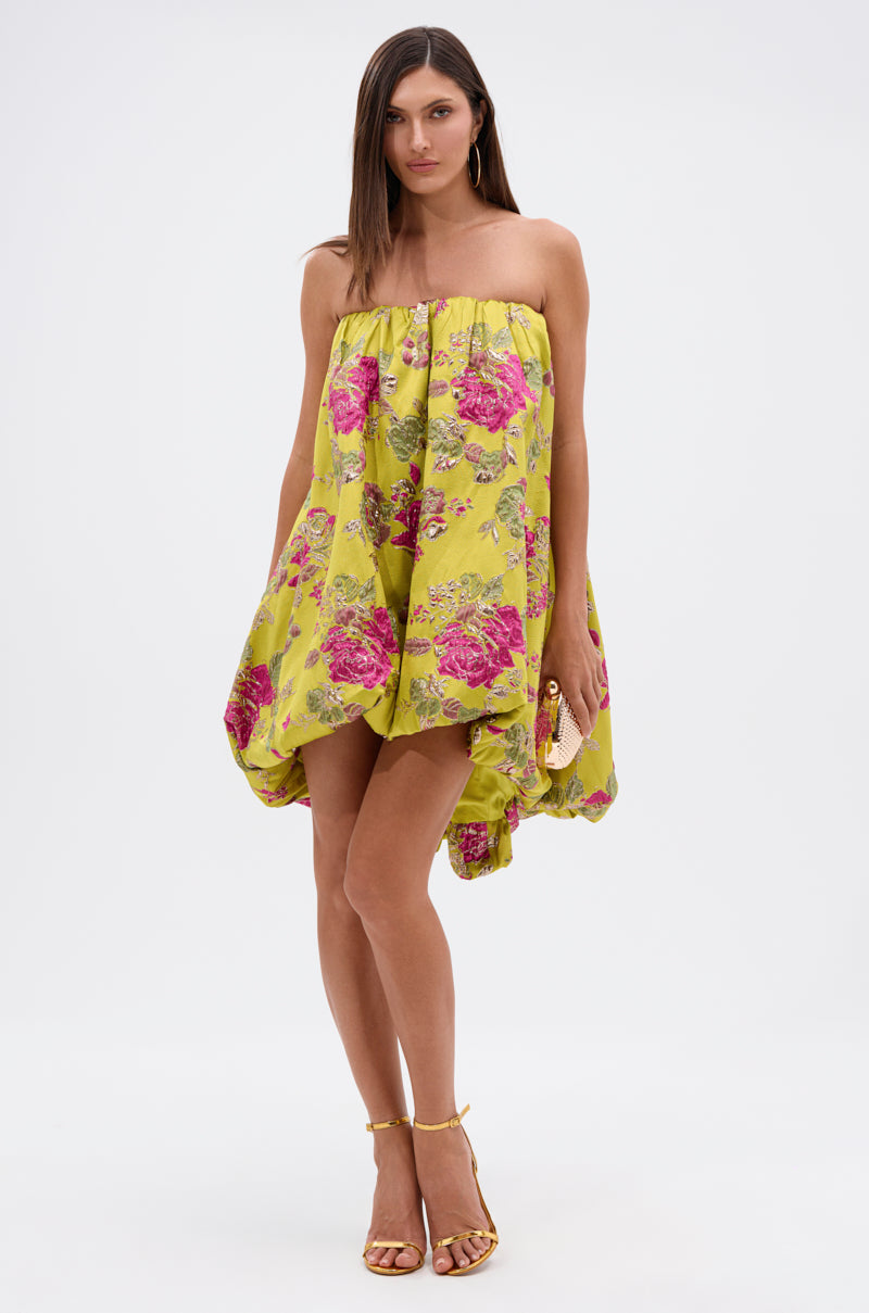 A woman models the LOVER GIRL BROCADE BUBBLE DRESS—couture-inspired, strapless, yellow with a pink floral pattern—paired with gold high-heeled sandals and a small gold clutch, set against a plain white background.