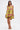 A woman models the LOVER GIRL BROCADE BUBBLE DRESS—couture-inspired, strapless, yellow with a pink floral pattern—paired with gold high-heeled sandals and a small gold clutch, set against a plain white background.