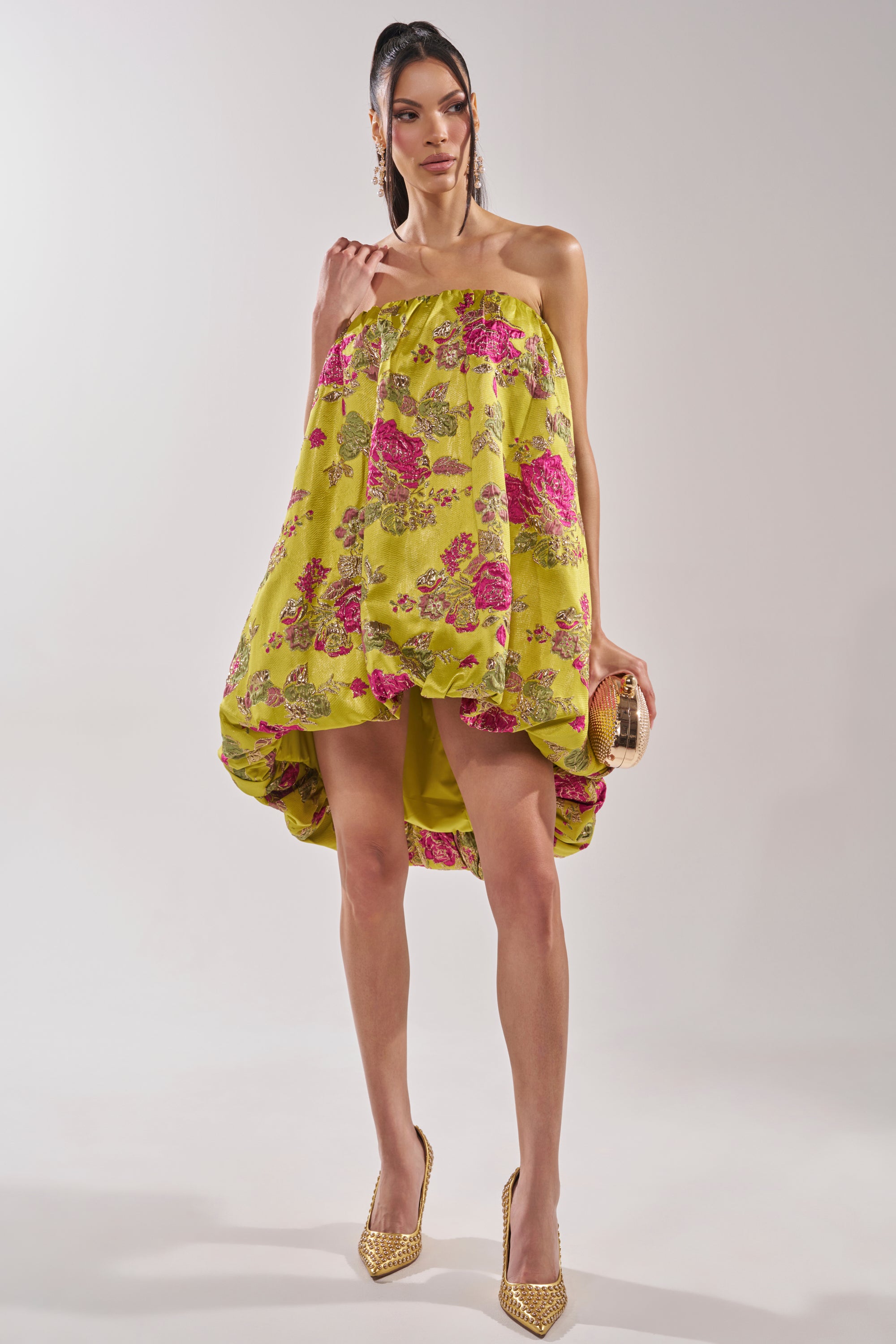 A woman stands confidently in the LOVER GIRL BROCADE BUBBLE DRESS, a yellow strapless mini with pink metallic floral embroidery. She holds a small clutch, wears gold studded heels, and styles her long hair in a sleek ponytail against a white background.