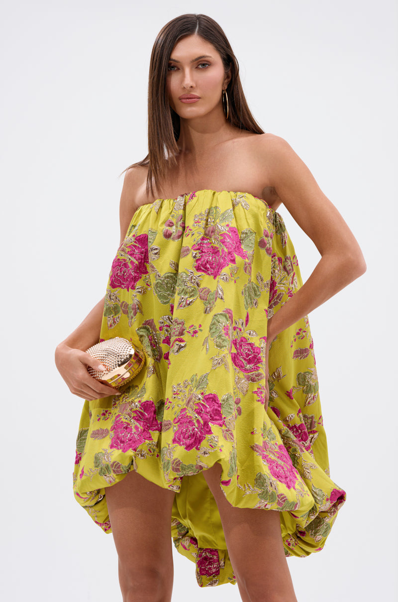 A woman wearing the LOVER GIRL BROCADE BUBBLE DRESS—strapless, yellow, with pink floral patterns—holds a gold clutch and looks at the camera against a plain light background.