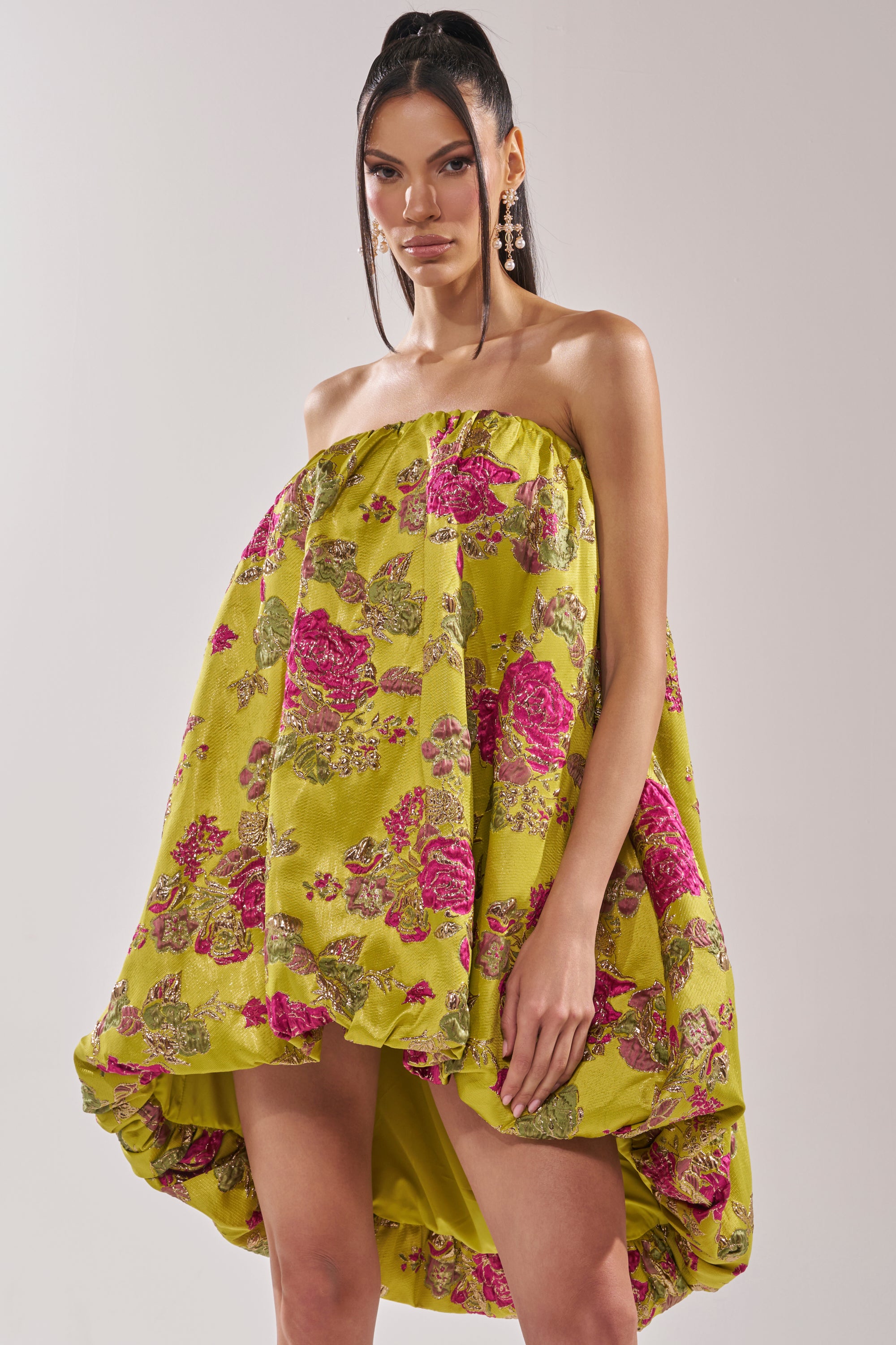 A woman models the LOVER GIRL BROCADE BUBBLE DRESS, a strapless mini with a voluminous high-low hem and pink and green metallic floral embroidery. She pairs it with a high ponytail, bold makeup, and statement earrings.