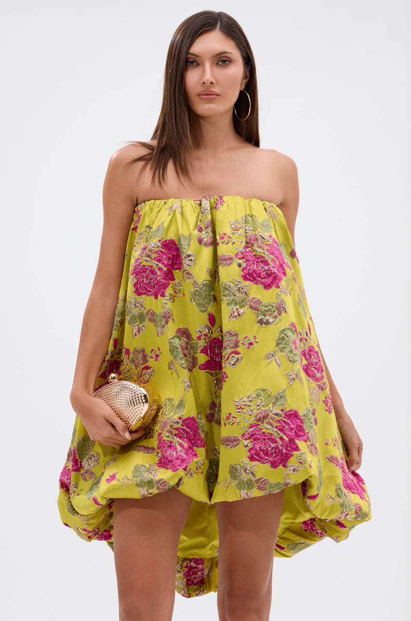 A woman with long brown hair models the LOVER GIRL BROCADE BUBBLE DRESS, featuring yellow brocade with pink and metallic floral details. She accessorizes with a gold clutch and large hoop earrings, standing before a white background.