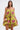 A woman with long brown hair models the LOVER GIRL BROCADE BUBBLE DRESS, featuring yellow brocade with pink and metallic floral details. She accessorizes with a gold clutch and large hoop earrings, standing before a white background.