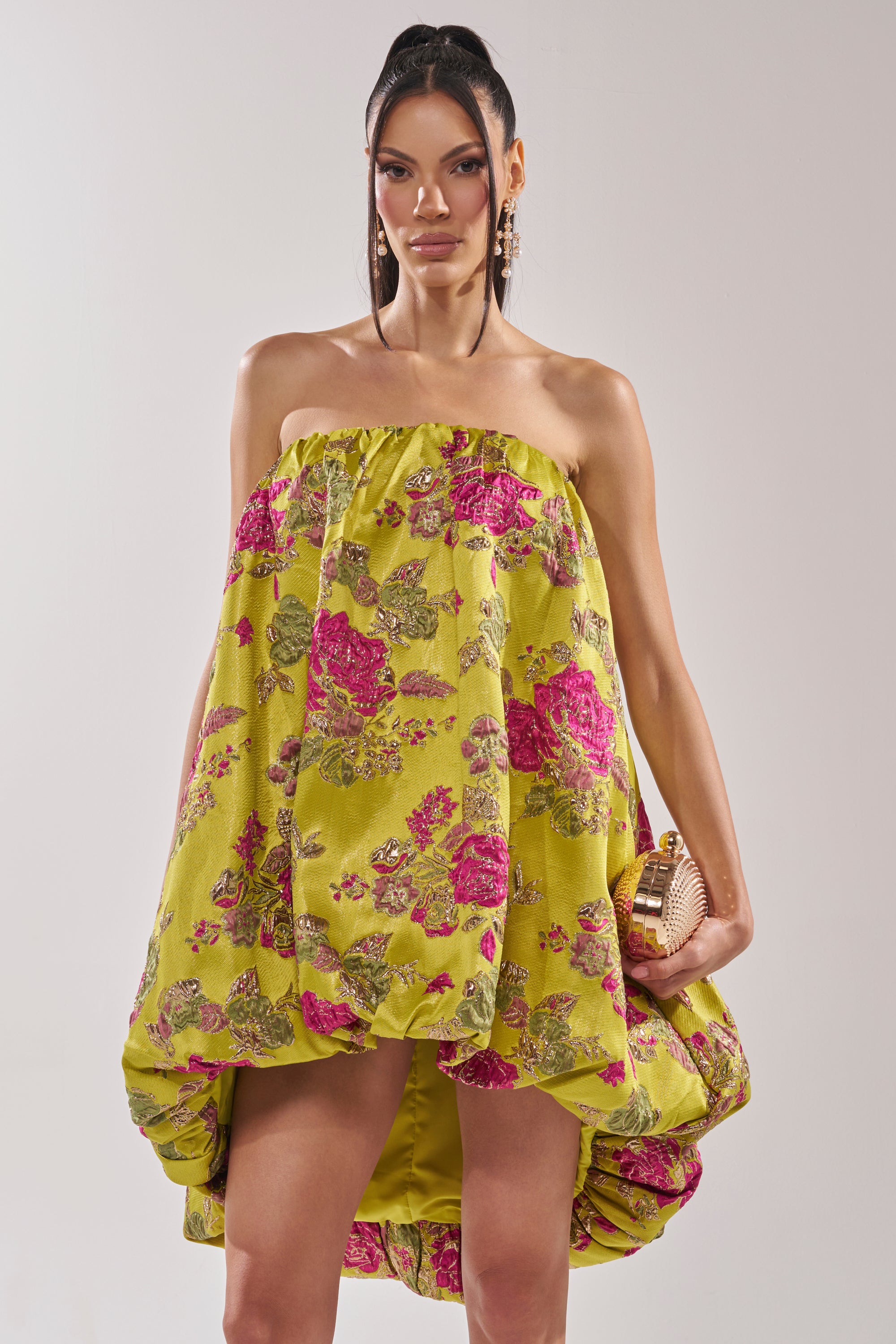 A woman models the LOVER GIRL BROCADE BUBBLE DRESS, a strapless yellow mini with pink floral patterns, paired with a gold textured clutch and statement earrings against a plain light background.