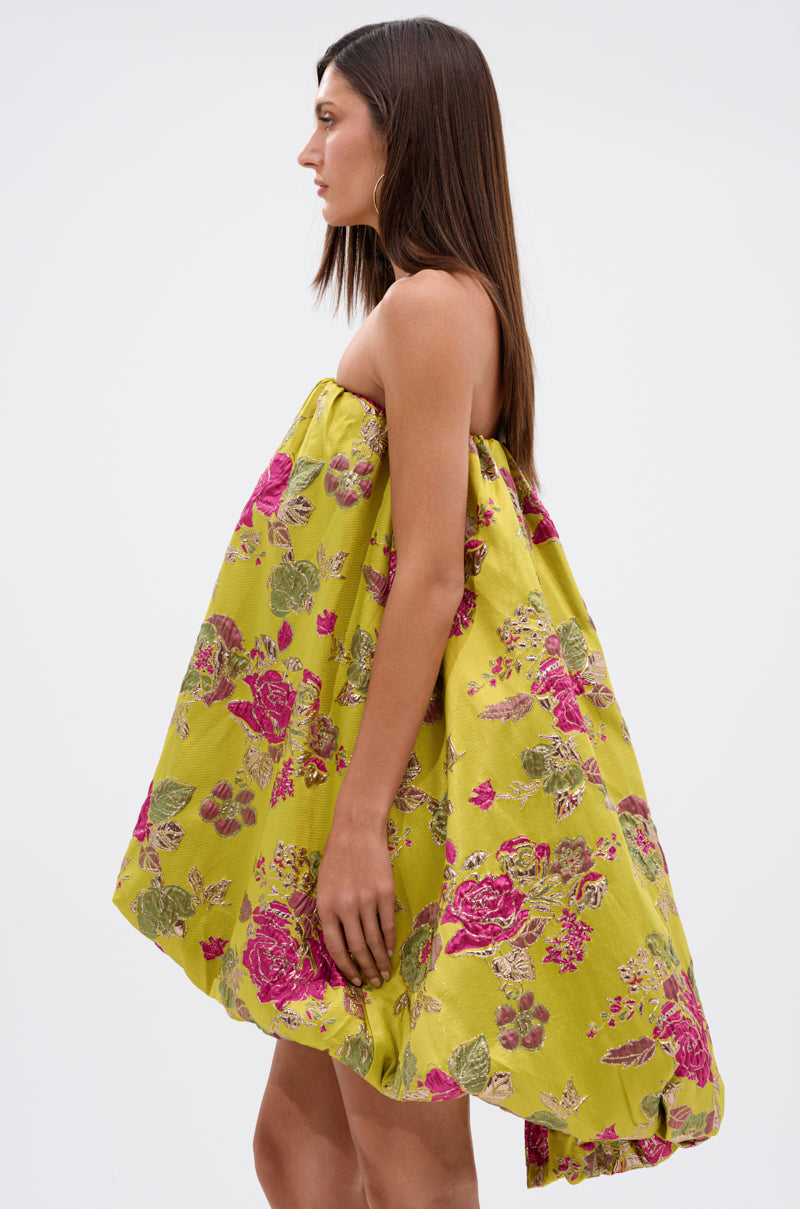 A woman in profile wears the LOVER GIRL BROCADE BUBBLE DRESS, a loose, strapless yellow-green brocade style with a pink and green floral pattern, standing out against a plain white background.