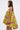 A woman in profile wears the LOVER GIRL BROCADE BUBBLE DRESS, a loose, strapless yellow-green brocade style with a pink and green floral pattern, standing out against a plain white background.
