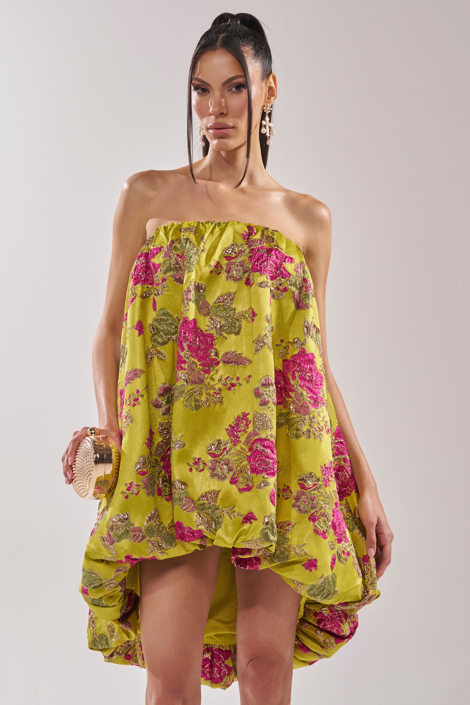 A woman wears the LOVER GIRL BROCADE BUBBLE DRESS, a strapless lime green mini with pink floral patterns and metallic embroidery. She holds a round gold clutch, wears statement earrings, and has her hair in a high ponytail.