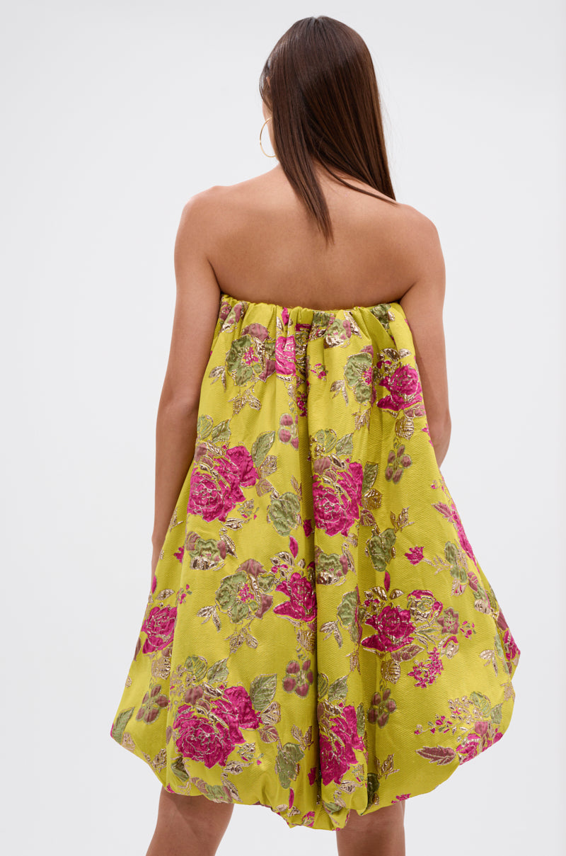 A woman with long, straight brown hair is shown from the back wearing the LOVER GIRL BROCADE BUBBLE DRESS, a couture-inspired strapless yellow dress with a pink and green floral pattern. The background is plain white.