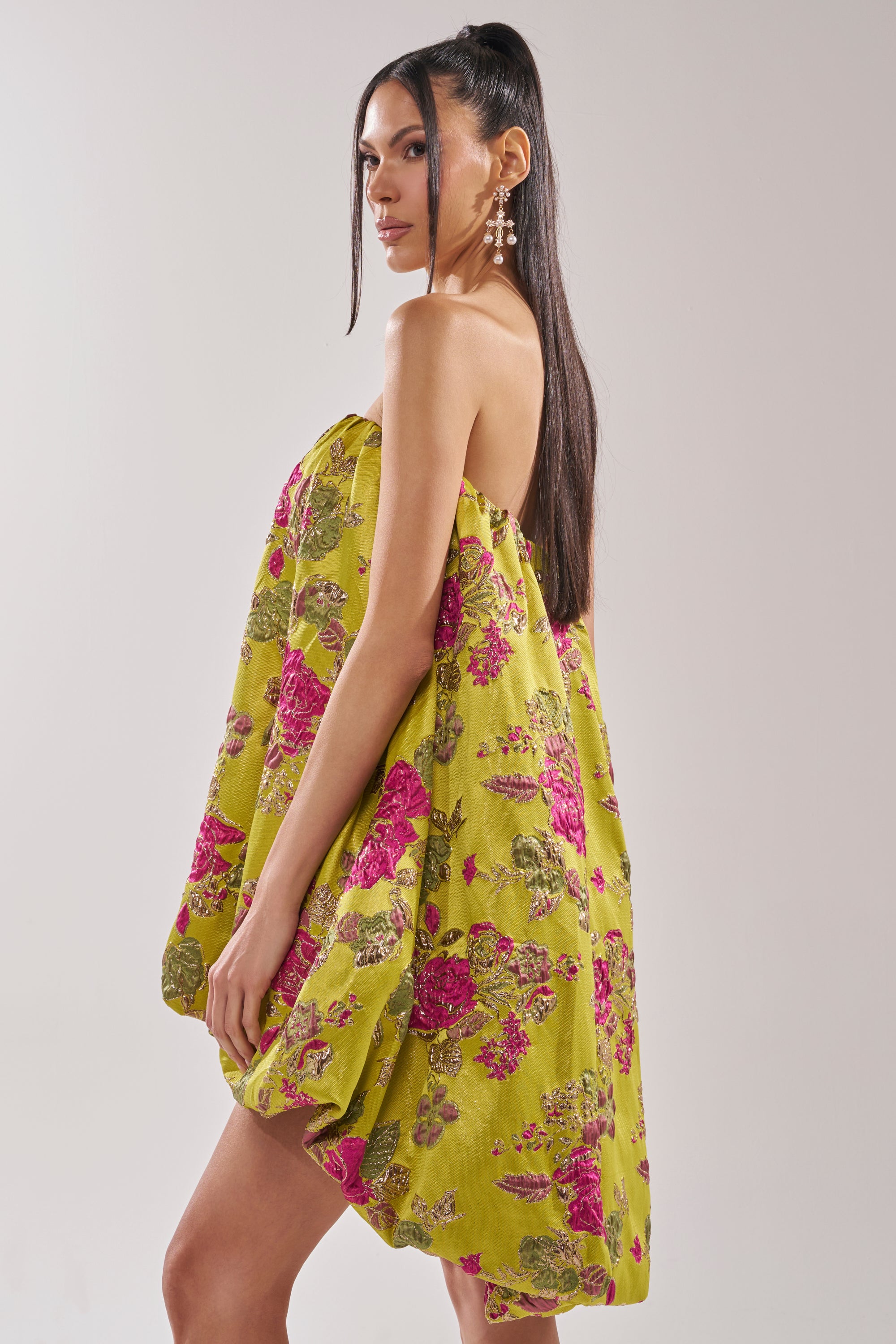 A woman with long dark hair models the LOVER GIRL BROCADE BUBBLE DRESS, a strapless mini dress with a yellow-green base and pink metallic floral embroidery, posing sideways and looking over her shoulder against a plain background.