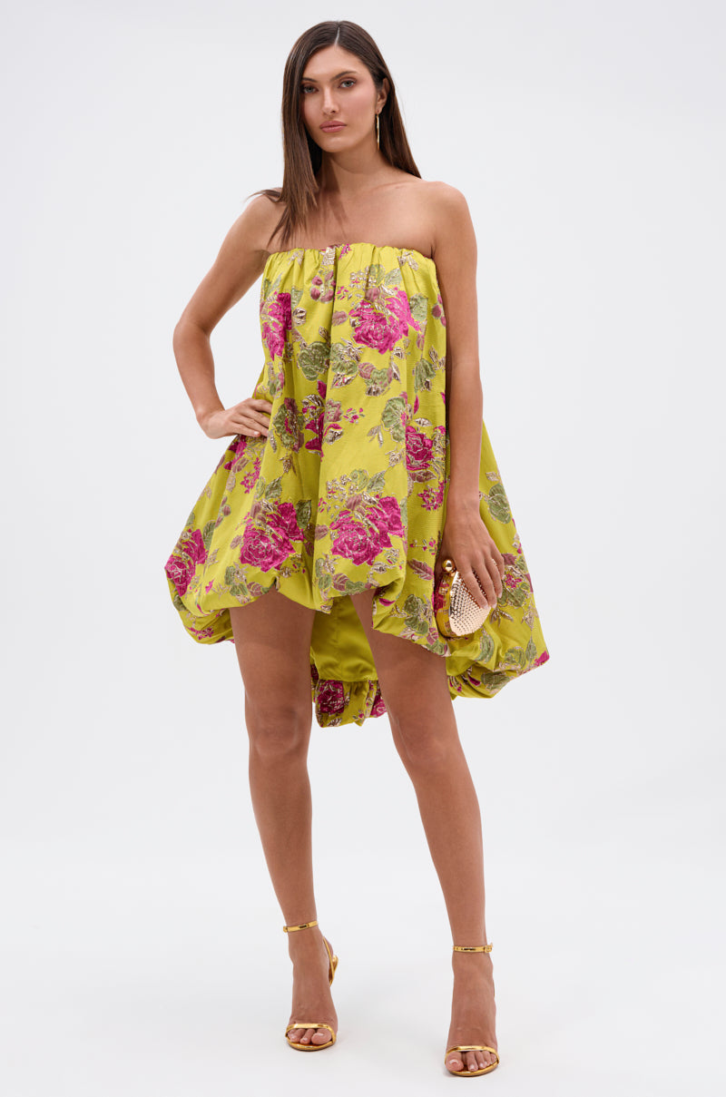 A woman in the LOVER GIRL BROCADE BUBBLE DRESS, a strapless yellow piece with pink and metallic floral details, stands against a white background holding a clutch, wearing yellow heels and her straight brown hair loose.