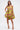 A woman in the LOVER GIRL BROCADE BUBBLE DRESS, a strapless yellow piece with pink and metallic floral details, stands against a white background holding a clutch, wearing yellow heels and her straight brown hair loose.