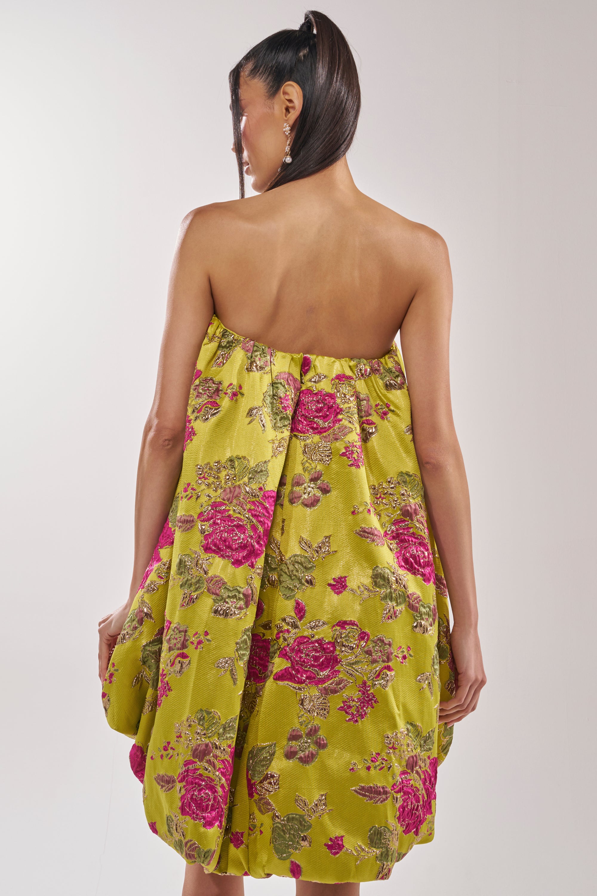 A woman with straight dark hair in a high ponytail faces away, wearing the LOVER GIRL BROCADE BUBBLE DRESS—a strapless mini dress with metallic pink and green floral embroidery, loose-fit silhouette, and a bubbly shape against a plain light backdrop.