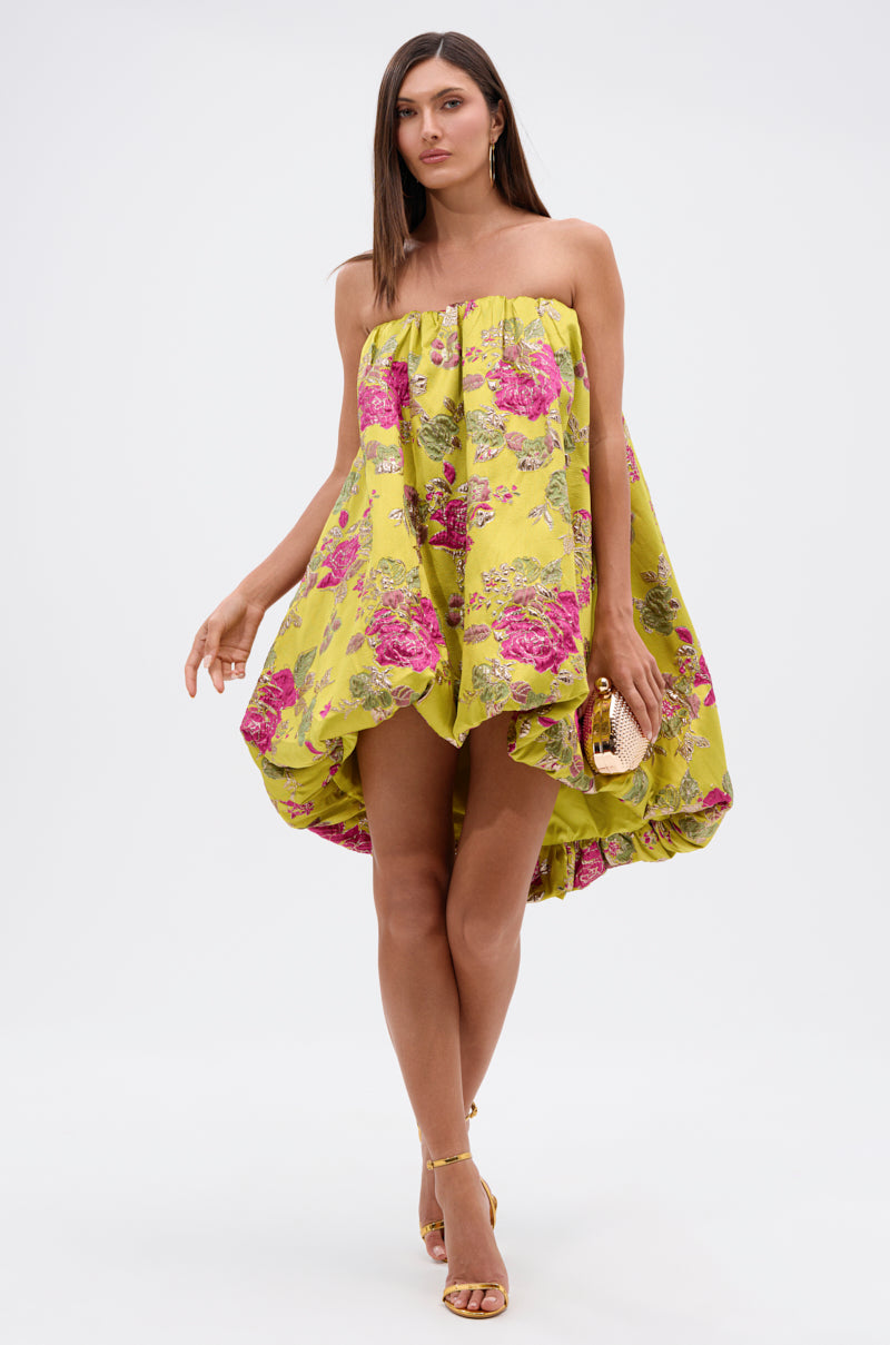 A woman in the LOVER GIRL BROCADE BUBBLE DRESS, featuring a strapless, loose-fit yellow design with pink and metallic floral accents, holds a small clutch. She wears yellow heels and stands against a plain white background.