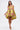 A woman in the LOVER GIRL BROCADE BUBBLE DRESS, featuring a strapless, loose-fit yellow design with pink and metallic floral accents, holds a small clutch. She wears yellow heels and stands against a plain white background.