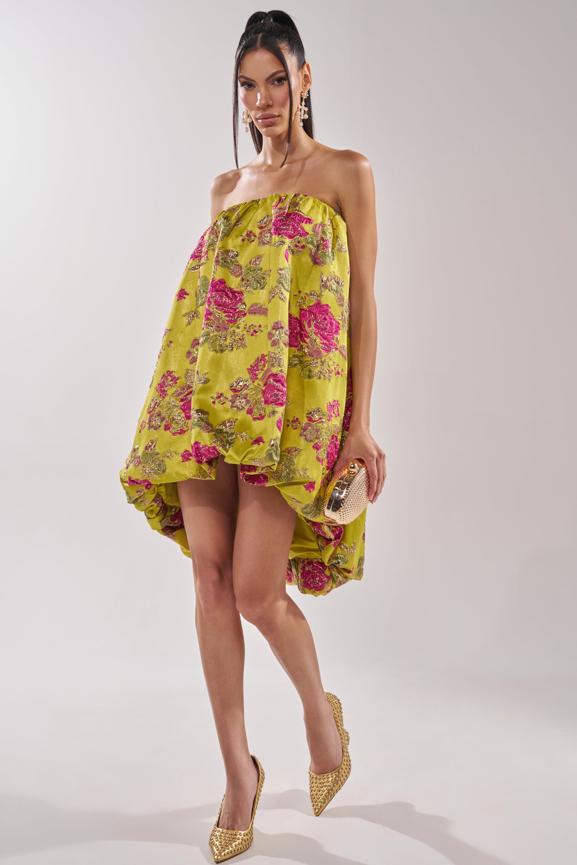 A woman poses in the LOVER GIRL BROCADE BUBBLE DRESS—strapless, with a high-low hemline and pink floral metallic embroidery. She accessorizes with a gold clutch, gold pointed heels, and statement earrings against a plain background.