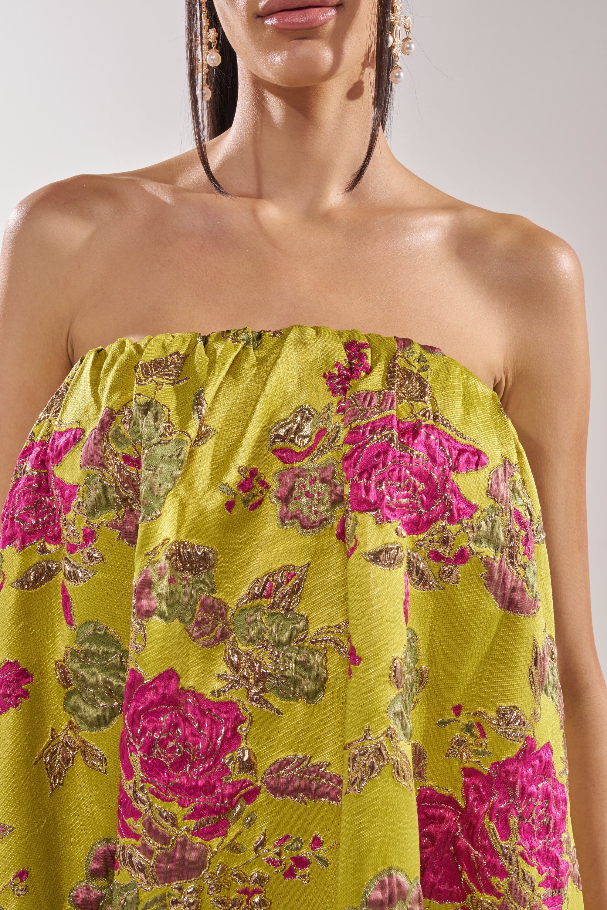 A woman models the LOVER GIRL BROCADE BUBBLE DRESS, a strapless yellow-green brocade dress with pink and green metallic floral embroidery, styled with dangling earrings. She is shown from shoulders to upper waist.