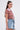 A young woman with long dark hair stands in profile against a plain light background, wearing large square earrings and the BEACHY BLISS CROCHET SHORT SLEEVE BUTTON DOWN with high-waisted denim shorts.