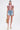 A woman faces forward in the BEACHY BLISS CROCHET SHORT SLEEVE BUTTON DOWN over a light pink top, high-waisted belted denim shorts, platform sandals, and holding a woven bag against a plain white background.