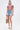 A woman stands against a white background wearing the Beachy Bliss Crochet Short Sleeve Button Down, high-waisted denim shorts, tan platform sandals, and holding a woven bag. Her hair is tied back and she sports large hoop earrings.