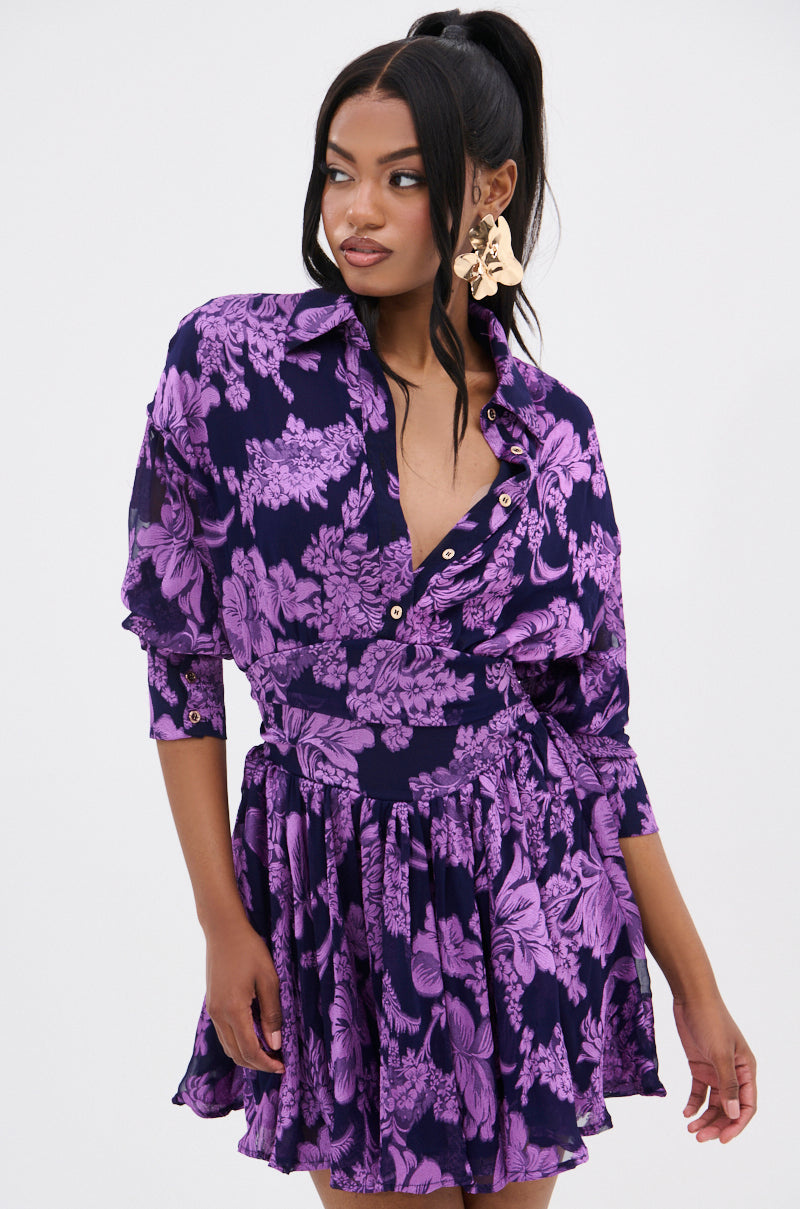 A woman poses in a WOMAN OF THE HOUR PRINTED MINI DRESS, showcasing a dark purple and lavender floral pattern. Her long, dark hair is in a high ponytail, complemented by large gold earrings against a plain white background.