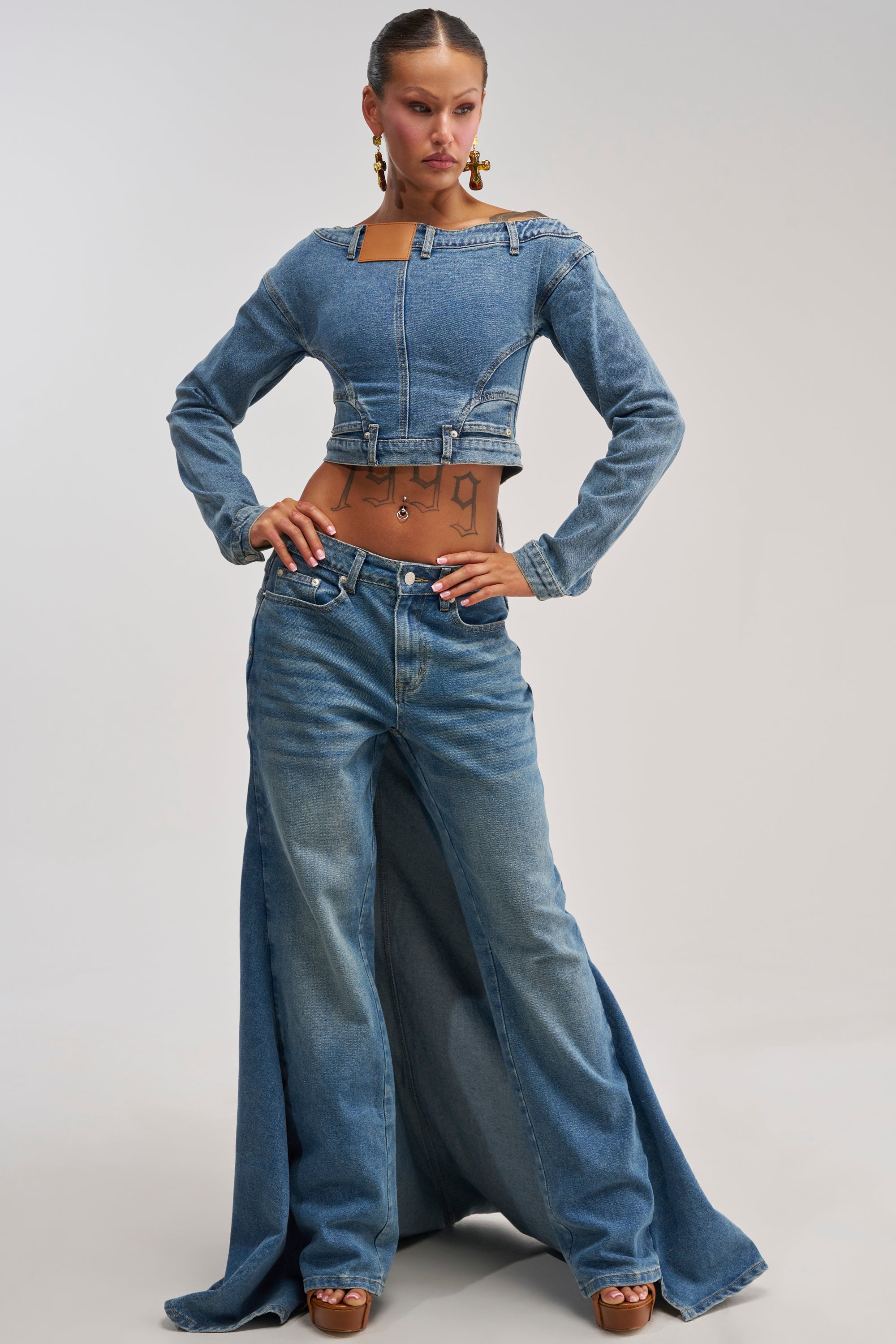 A woman poses in the SHAKE IT OFF DENIM PANT WITH SKIRT ATTACHED, featuring wide-leg, floor-length denim with a high waist. She styles the look with heels, gold earrings, and a sleek ponytail, revealing a torso tattoo.