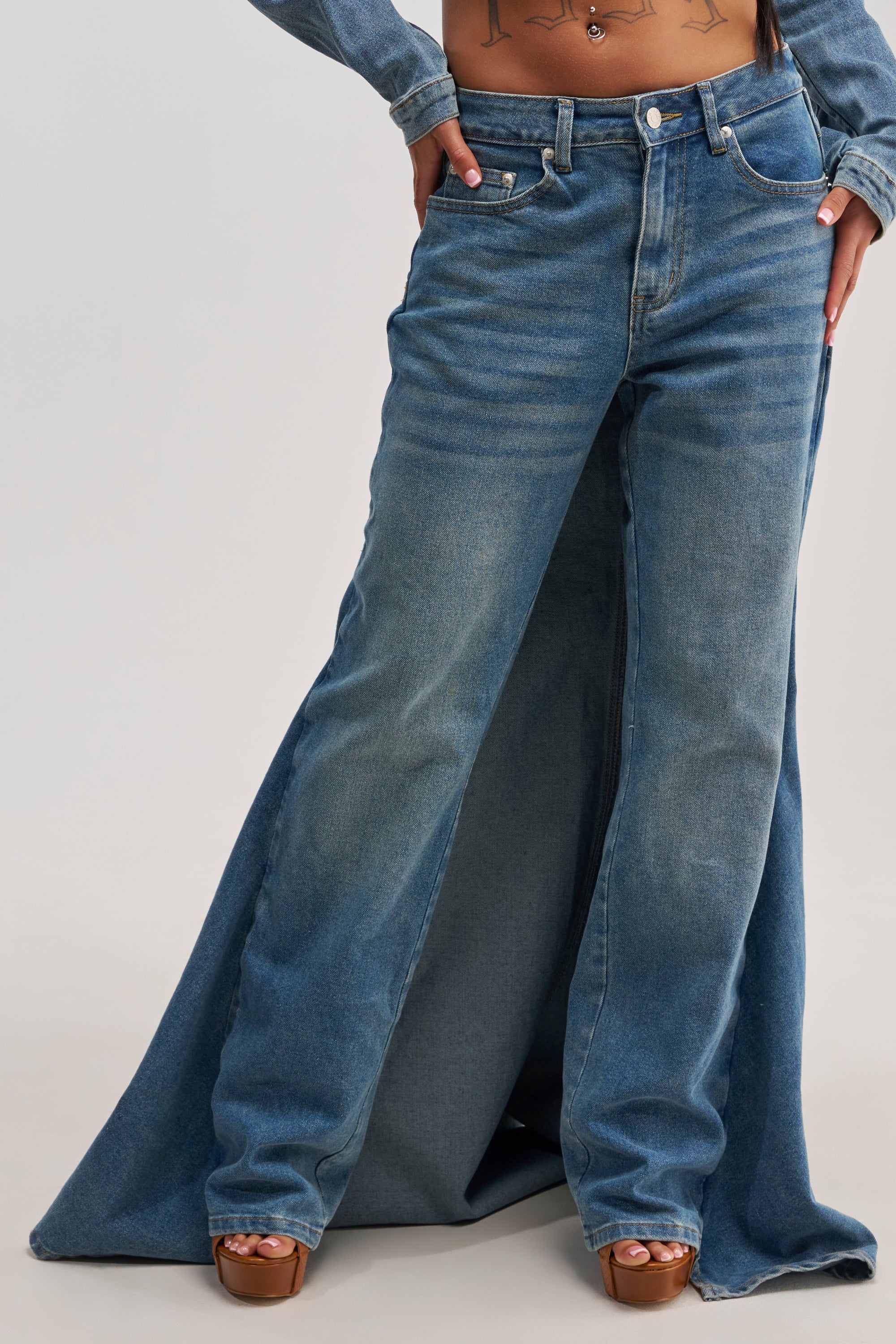 A person wearing the SHAKE IT OFF DENIM PANT WITH SKIRT ATTACHED, featuring wide-leg blue jeans made from stretch denim that flare dramatically at the bottom and cover their shoes. Their hands rest on their hips, with only the lower half of their body visible.