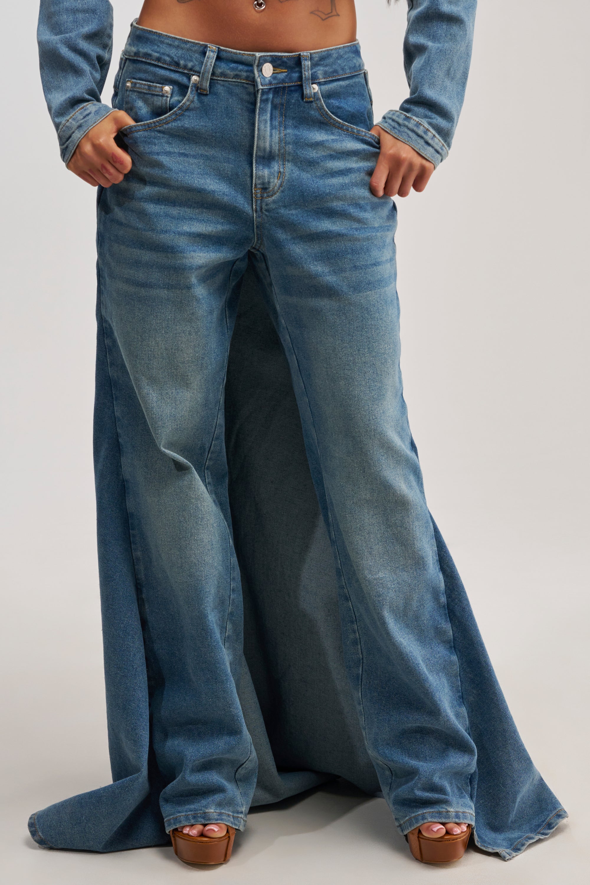 A person wearing the SHAKE IT OFF DENIM PANT WITH SKIRT ATTACHED, featuring wide-leg, high-waisted stretch denim and exaggerated flared panels, stands with hands in pockets and toes peeking out from open-toe brown shoes.