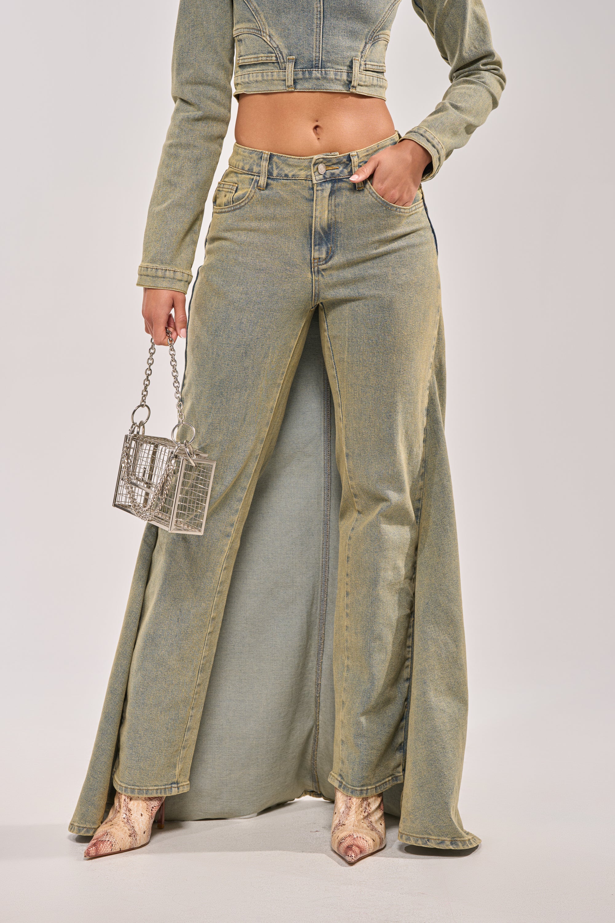 A woman wears the BIG STEPPER DENIM PANT, a high-waisted acid wash maxi skirt with a front slit, styled with a matching cropped denim jacket, snakeskin pointed-toe boots, and a small metallic handbag. 0