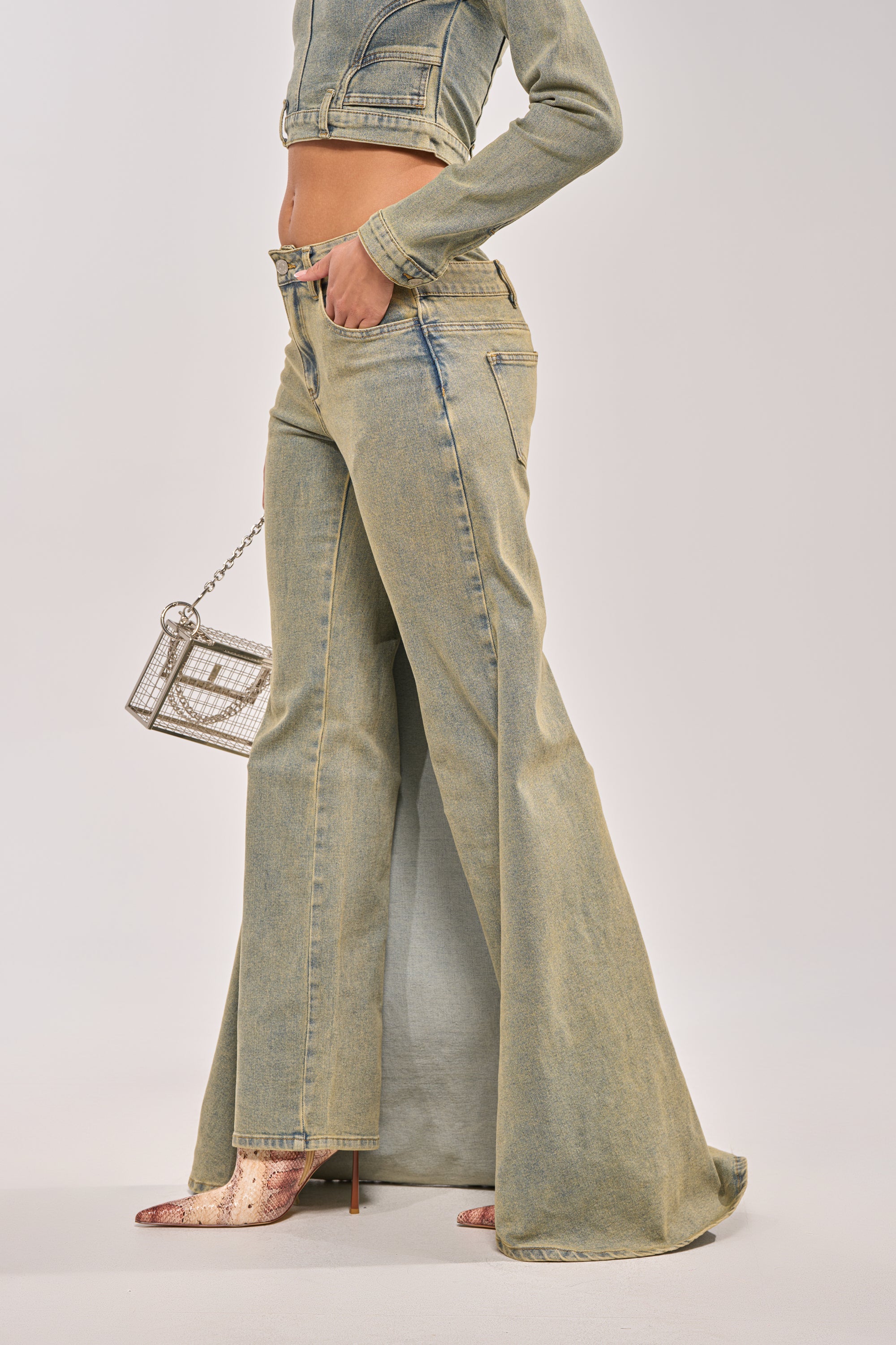 A person wears the BIG STEPPER DENIM PANT styled with a cropped denim jacket, metallic pointed-toe heels, and carries a small rectangular chain-strap handbag. The background is plain and light-colored. 2