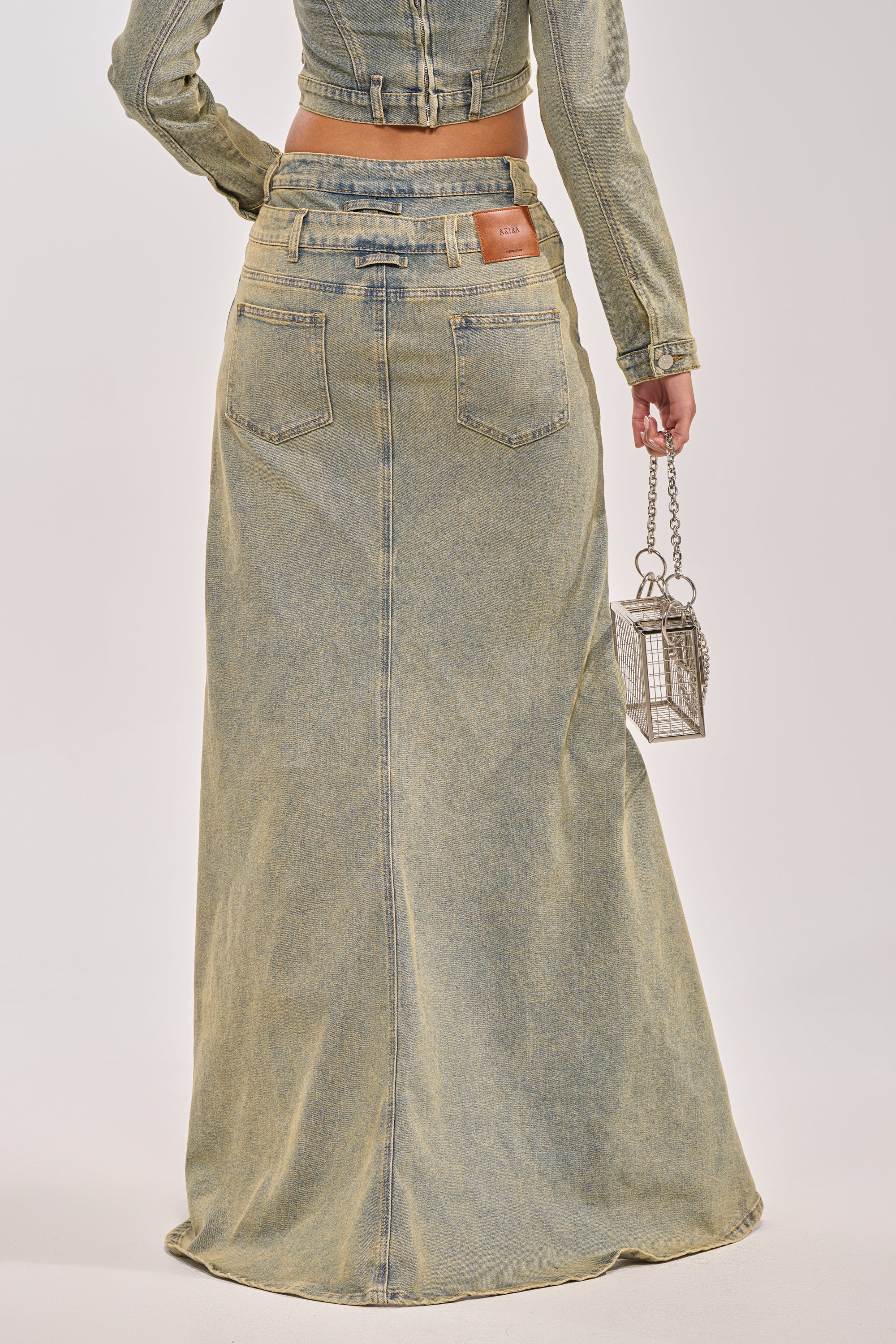 A person stands facing away, wearing the BIG STEPPER DENIM PANT—a long, light-wash denim style—paired with a matching cropped jacket and metallic handbag, showcasing statement streetwear with effortless cool. 3