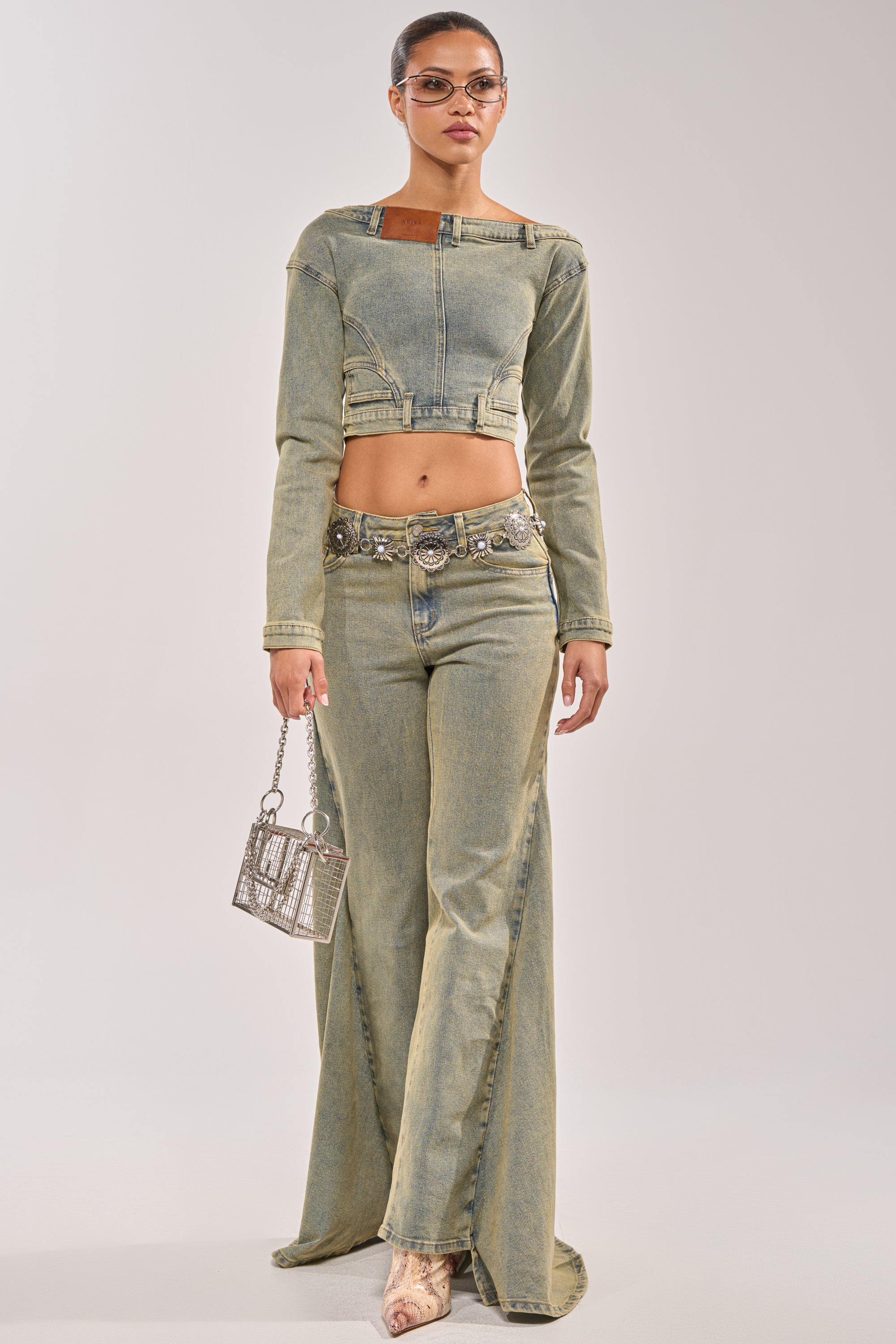 A woman stands against a plain light background in matching acid wash, light green BIG STEPPER DENIM PANTS and a cropped denim jacket, styled with a silver belt, sunglasses, and a metallic mini handbag for an effortlessly bold streetwear look. 4