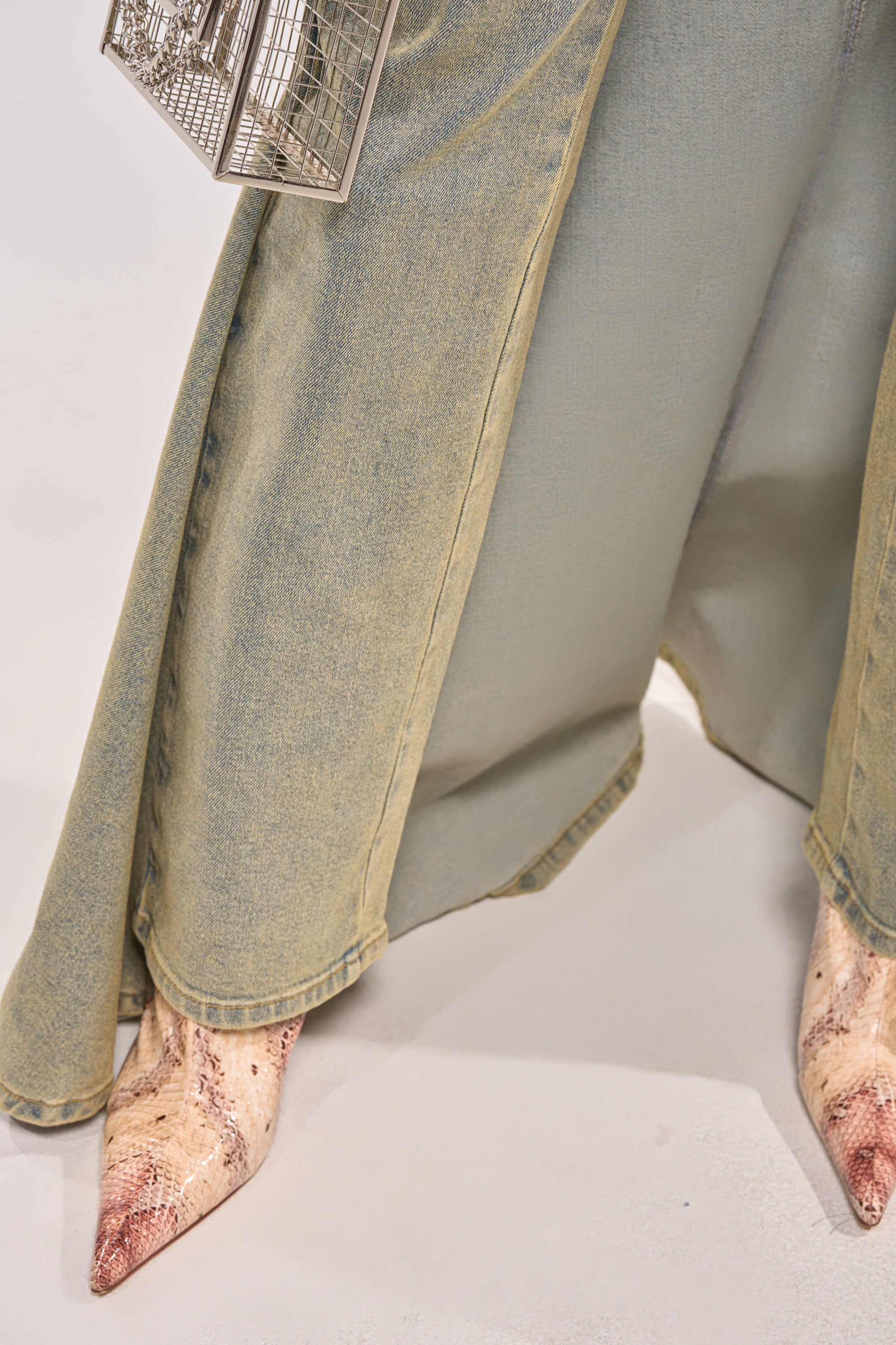 A person wears the BIG STEPPER DENIM PANT in light-wash denim with pointed snake-print boots and holds a small silver geometric handbag, standing on a light surface. 6