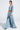 Confidently standing in a stylish 2-in-1 ensemble, a person wears an AKIRA Label Shake It Off Denim Pant with Skirt Attached and a long-sleeve off-shoulder cropped top, completed with chunky heels and earrings, against a plain white background.
