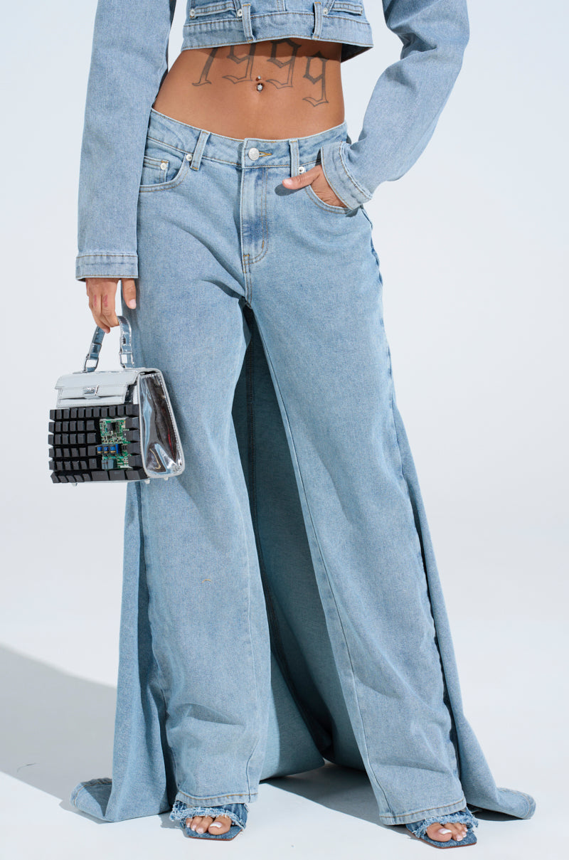 Wearing the SHAKE IT OFF DENIM PANT WITH SKIRT ATTACHED and a cropped denim jacket, a person shows off a tattoo on their midriff while holding a shiny handbag. The jeans have an ultra-wide flare from the waist and boast a chic denim skirt overlay, enhancing their eye-catching 2-in-1 design.