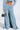 Wearing the SHAKE IT OFF DENIM PANT WITH SKIRT ATTACHED and a cropped denim jacket, a person shows off a tattoo on their midriff while holding a shiny handbag. The jeans have an ultra-wide flare from the waist and boast a chic denim skirt overlay, enhancing their eye-catching 2-in-1 design.