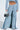 Person wearing SHAKE IT OFF DENIM PANT WITH SKIRT ATTACHED, featuring a light blue, wide-leg design and a matching cropped jacket. A small metallic handbag complements the tattooed midriff, while denim-strapped open-toe heels stand out against a plain white background.