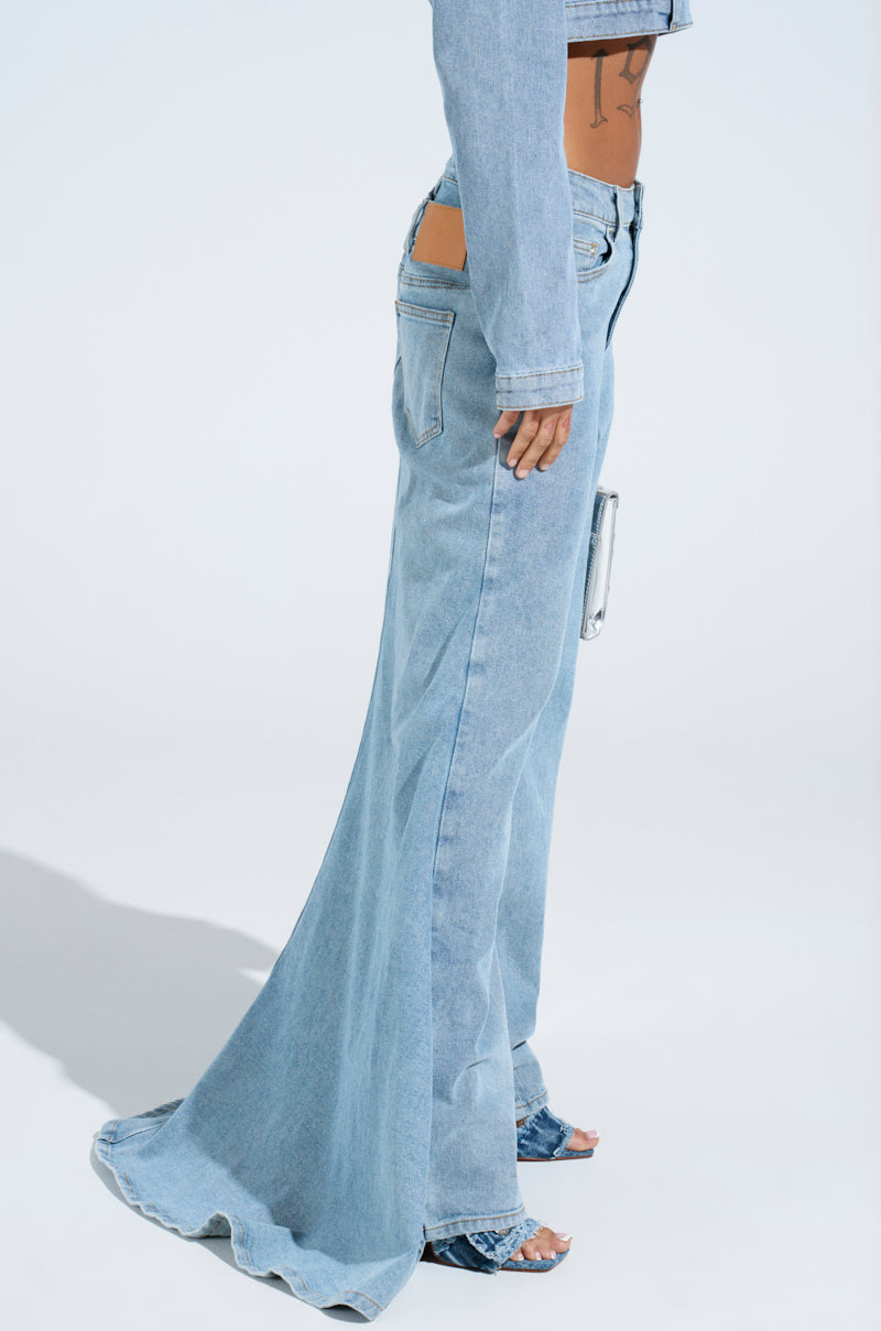 A person models the AKIRA Label Shake It Off Denim Pant with Skirt Attached, showcasing exaggerated flares and stylish back pocket cut-outs, paired with a matching cropped denim jacket against a plain backdrop, completing the chic 2-in-1 design.