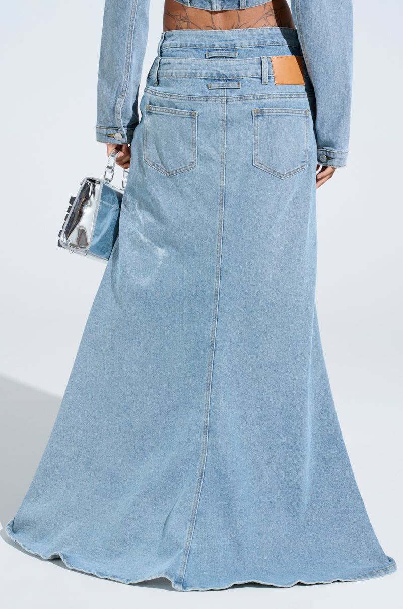 Modeling the SHAKE IT OFF DENIM PANT WITH SKIRT ATTACHED resembling stacked jeans, a person carries a small silver handbag and wears a matching denim jacket against a plain background.