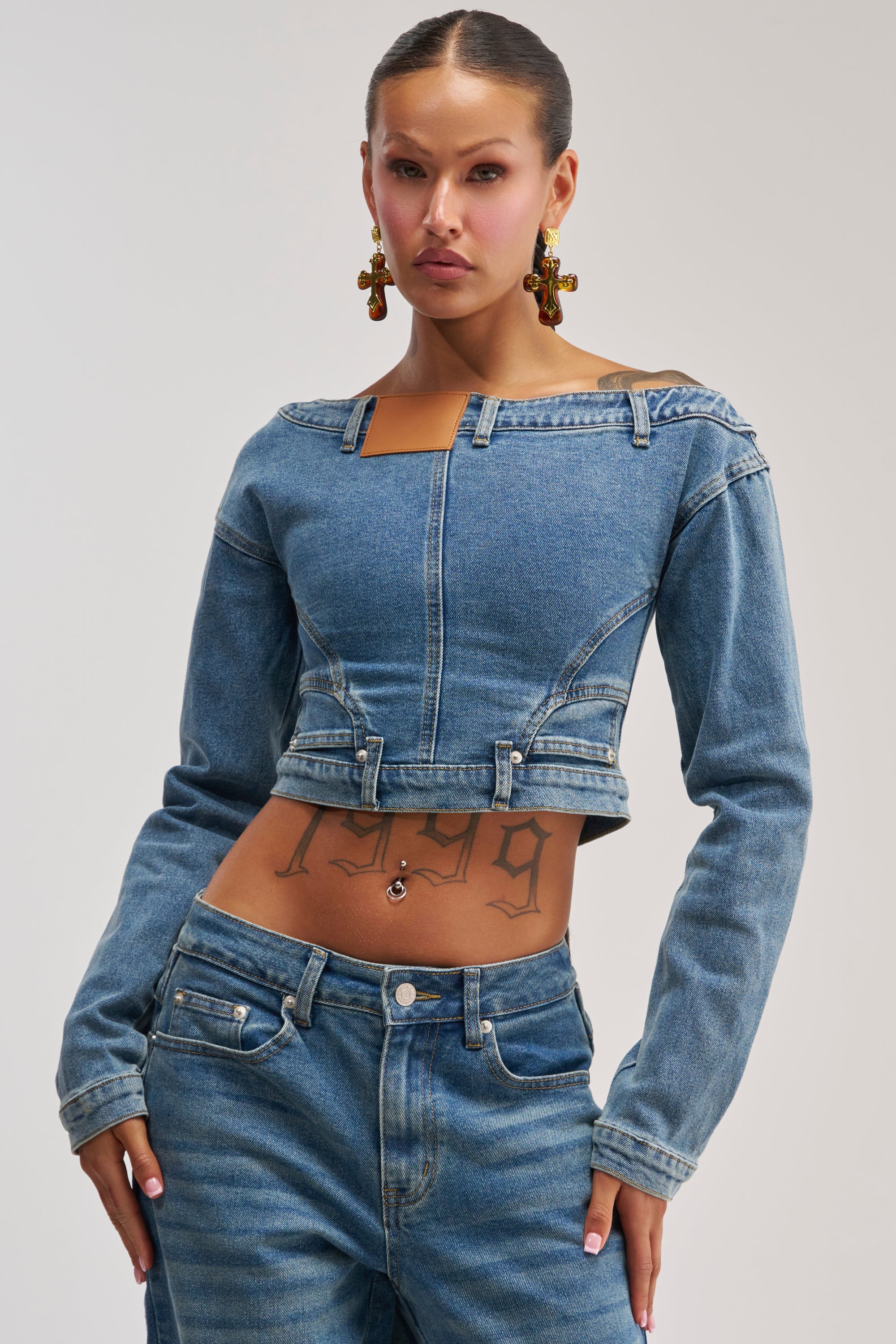 Wearing the SHAKE IT OFF DENIM OFF THE SHOULDER TOP, a woman styled in streetwear reveals a visible "1999" tattoo on her stomach and statement earrings as she poses confidently against a plain background.