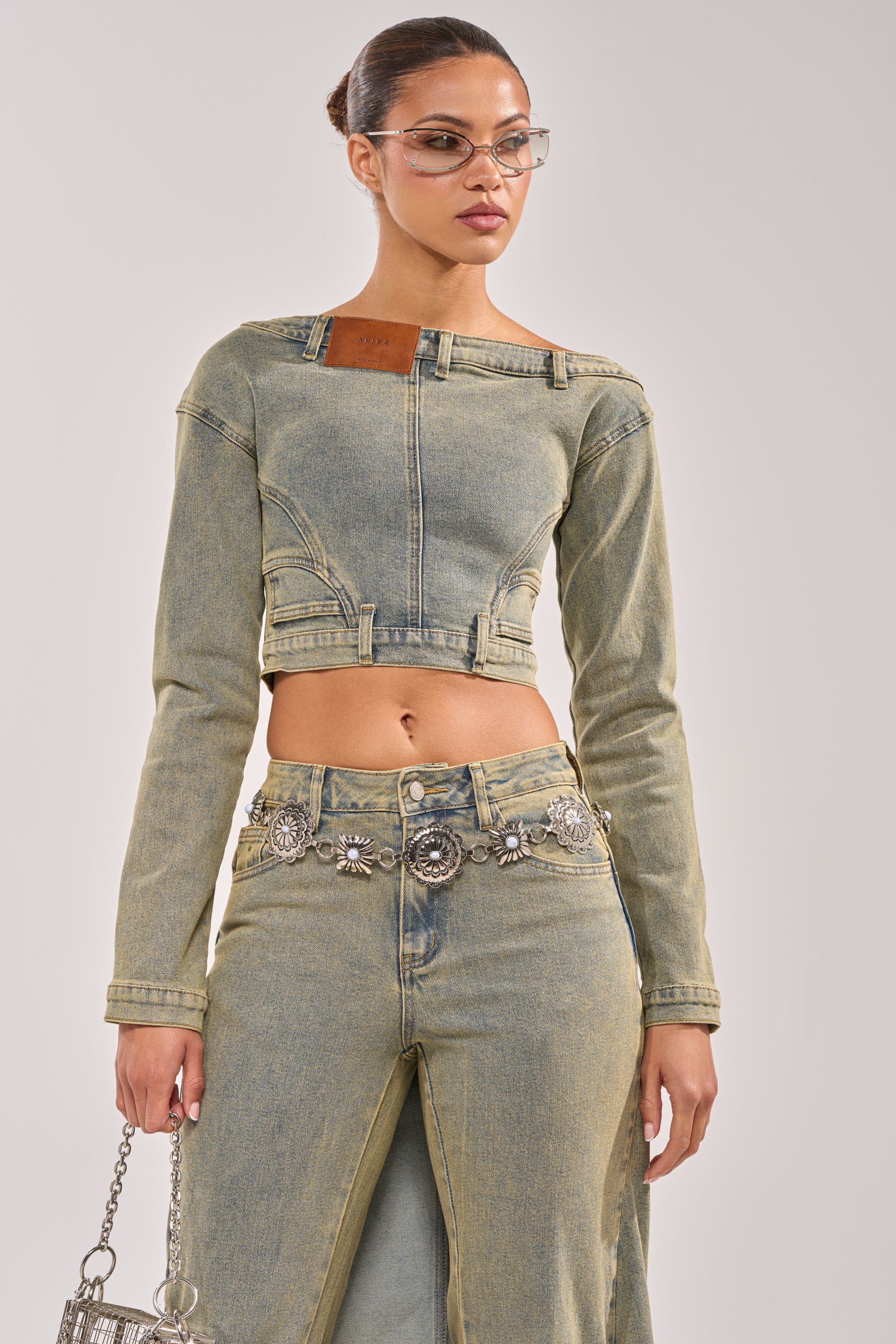 A woman wears the BIG STEPPER DENIM BLOUSE with a high-waisted denim skirt featuring a front slit, accessorized with a silver floral belt, glasses, and silver handbag, styled with streetwear vibes against a plain light background.