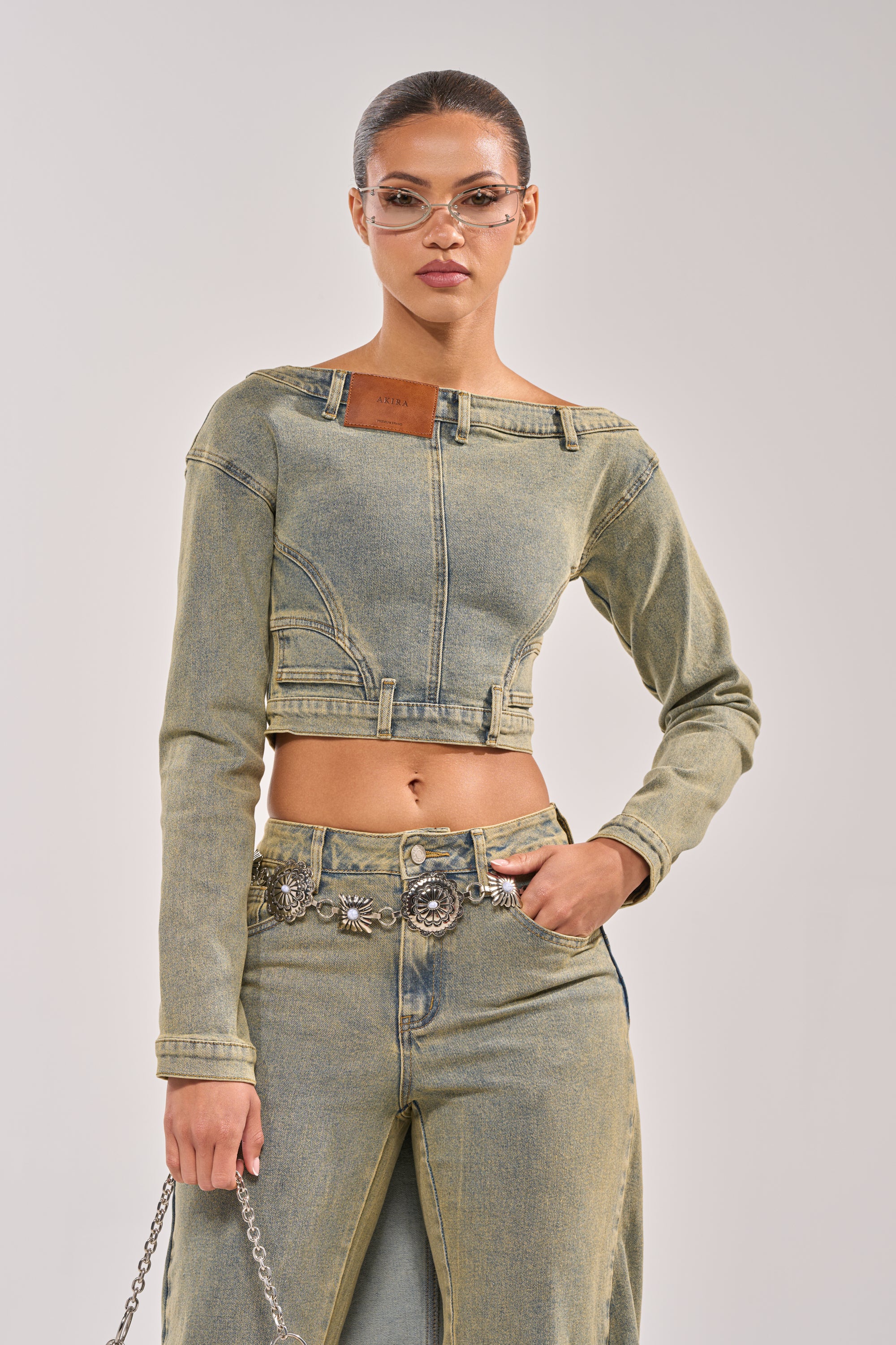 A woman wears the BIG STEPPER DENIM BLOUSE, paired with high-waisted jeans featuring a front slit, a silver belt, clear cat-eye glasses, and a chain strap bag, standing against a plain light background.
