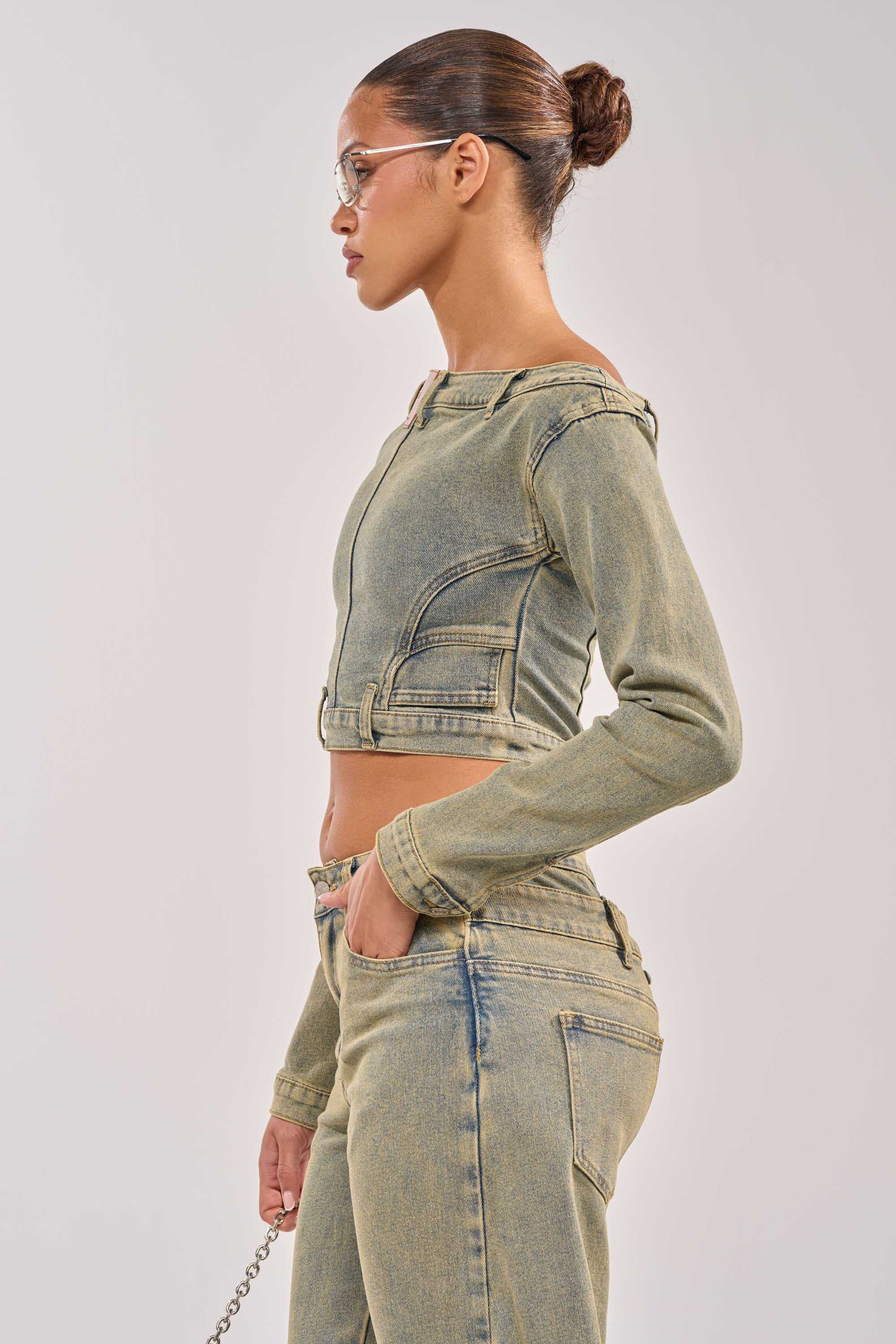 A woman in profile wears the BIG STEPPER DENIM BLOUSE and matching jeans with streetwear flair, her hair in a neat bun, one hand in her pocket, and clear-framed glasses, set against a plain background.
