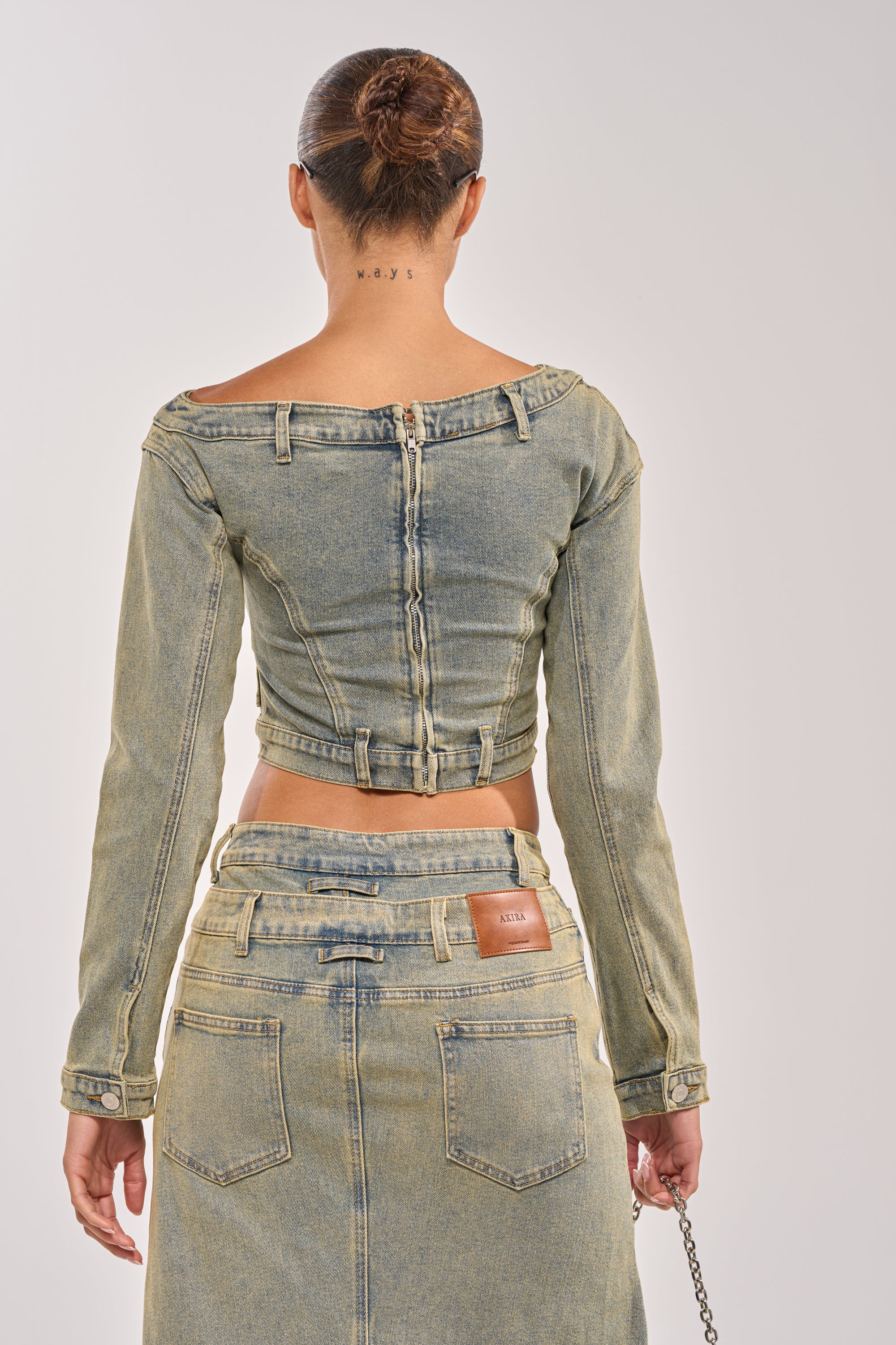 Seen from behind, a person with hair in a bun wears the BIG STEPPER DENIM BLOUSE off-shoulder, styled as streetwear with visible seams, paired with a matching high-waisted denim skirt featuring a brown patch above the back pocket.