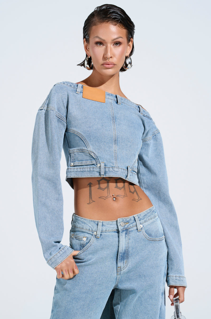 Person confidently posing in a SHAKE IT OFF DENIM OFF THE SHOULDER TOP by AKIRA Label. The light blue denim jacket, with its unique cut-out shoulder design and beige patch, complements their jeans. Tattoos adorn their torso as they hold an item against a light blue background.