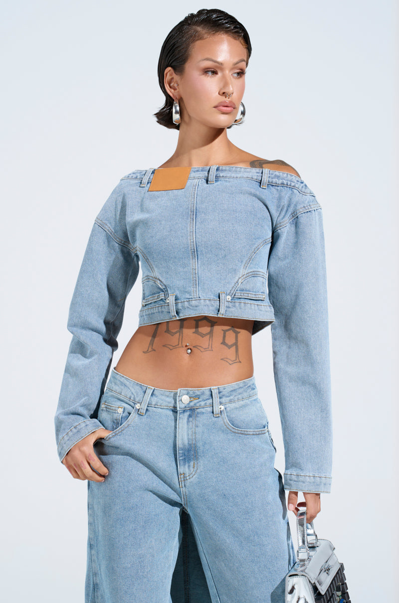 The person radiates confidence in a SHAKE IT OFF DENIM OFF THE SHOULDER TOP and jeans, with a metallic handbag. A "1999" tattoo decorates their stomach, and their sleek, short hair embodies the edgy spirit of the AKIRA Label as they glance sideways.