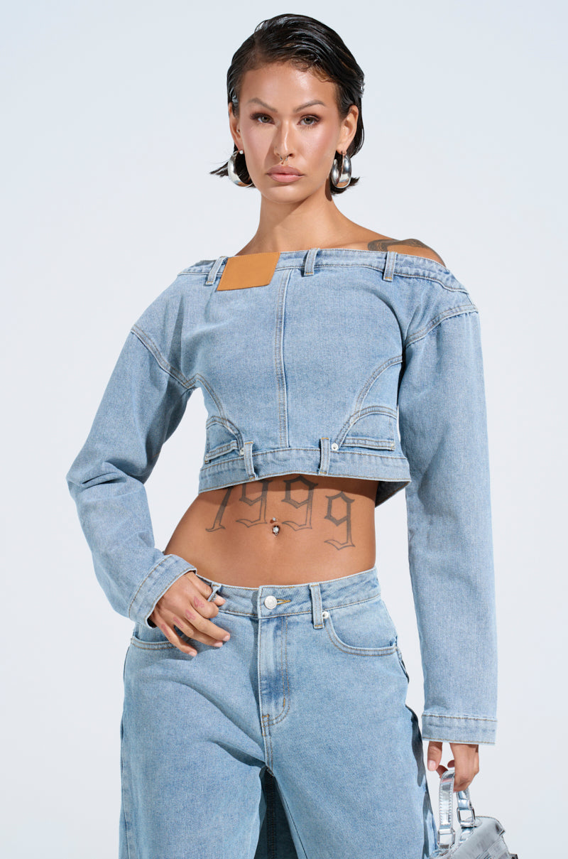 A person with short dark hair wears the SHAKE IT OFF DENIM OFF THE SHOULDER TOP with matching medium wash jeans. Tattoos decorate their stomach as they hold a small silver handbag featuring a metallic back zipper closure, set against a plain light background.