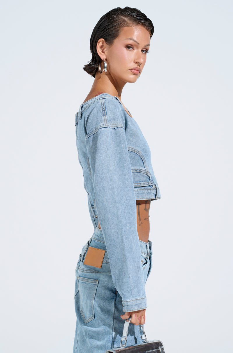 A person with short dark hair stands against a light background, wearing the SHAKE IT OFF DENIM OFF THE SHOULDER TOP with medium wash cotton denim pants. They hold a small handbag and look to the side, their style subtly accented by a metallic back zipper closure.