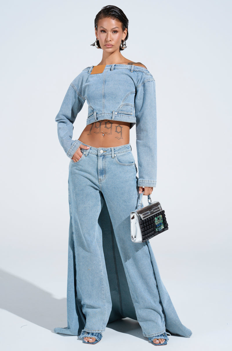 In a Shake It Off Denim off-the-shoulder top, someone confidently poses with a small bag against a simple background. Their short, slicked hair complements the AKIRA Label style, paired with wide-leg pants and open-toed shoes.