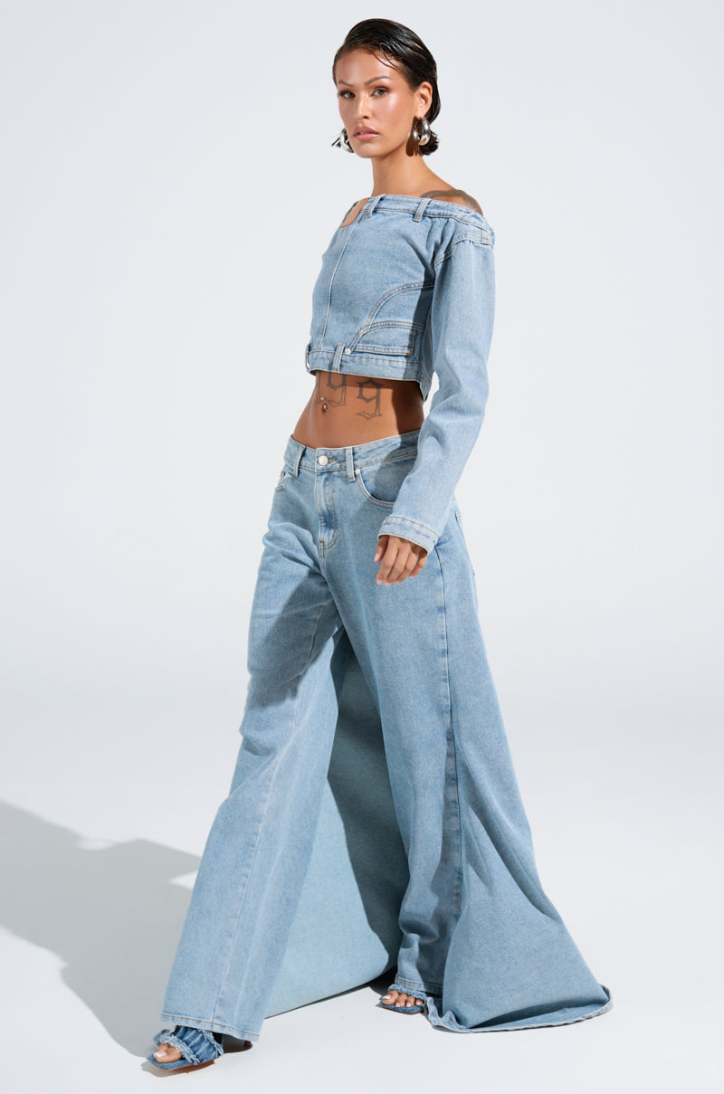 A person stands confidently in the AKIRA Label light blue SHAKE IT OFF DENIM OFF THE SHOULDER TOP and wide-leg jeans. Their slicked-back hair and statement earrings add flair. A plain white studio sets the scene, with their shadow elegantly cast on the floor.