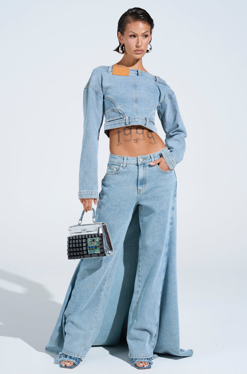 Confidently posing in a trendy Shake It Off Denim outfit, they sport an AKIRA Label off-the-shoulder top with wide-leg jeans. A chic handbag and minimal jewelry complete the look against a light background.